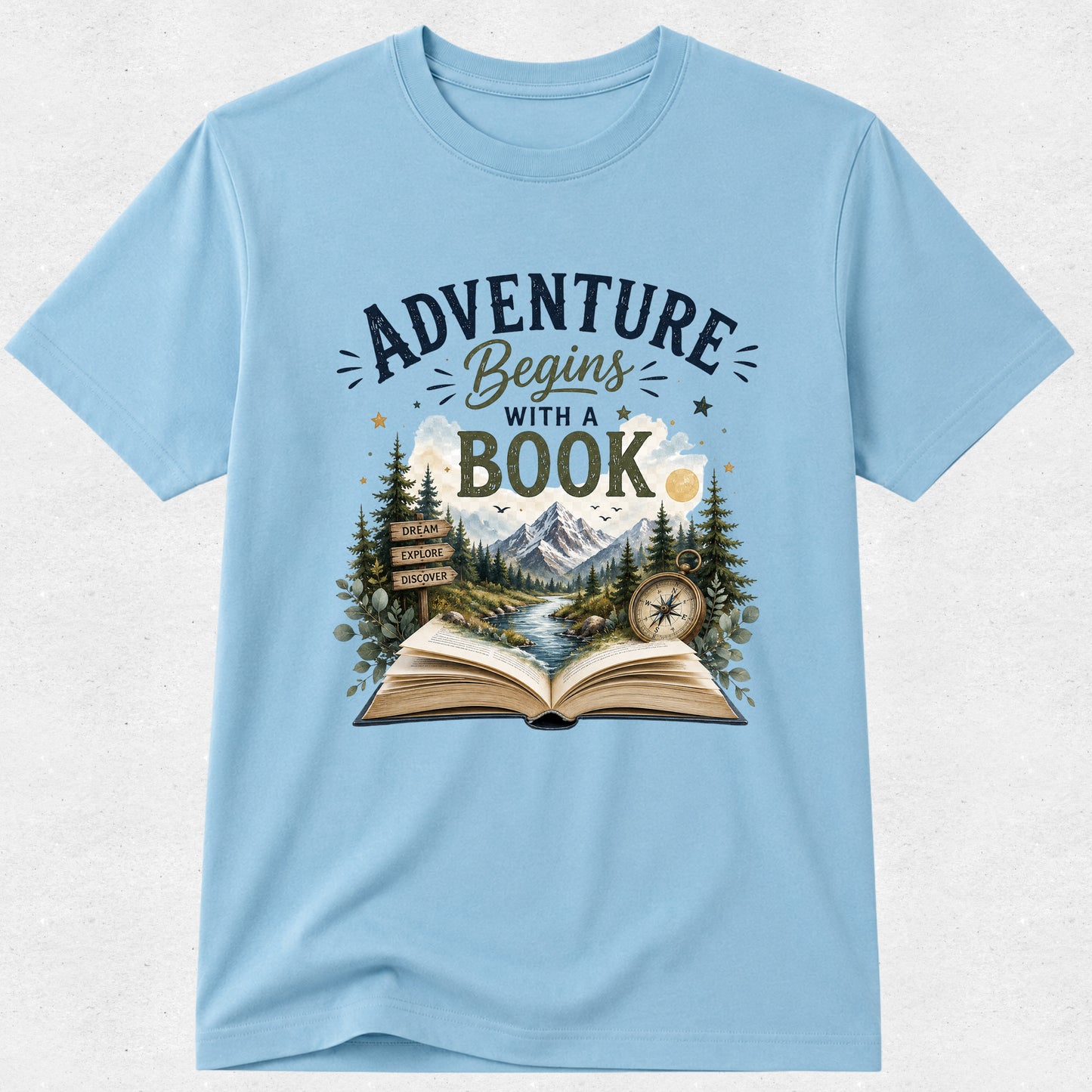 A light blue short-sleeve crewneck t-shirt featuring a graphic print on the front with the text 'ADVENTURE Begins WITH A BOOK' above an illustration of an open book, mountains, pine trees, a river, and a compass.