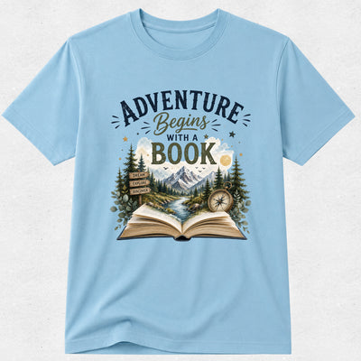 A light blue short-sleeve crewneck t-shirt featuring a graphic print on the front with the text 'ADVENTURE Begins WITH A BOOK' above an illustration of an open book, mountains, pine trees, a river, and a compass.