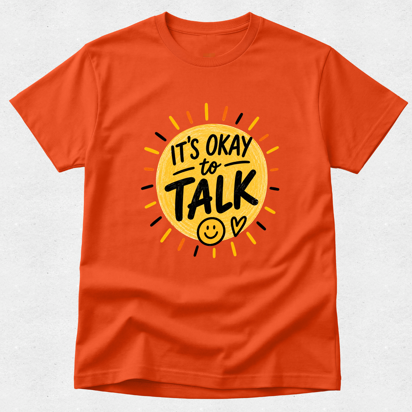 An orange short-sleeve crewneck t-shirt featuring a graphic design of a yellow sun with the text 'IT'S OKAY to TALK' and a smiley face, displayed on a light gray textured background.
