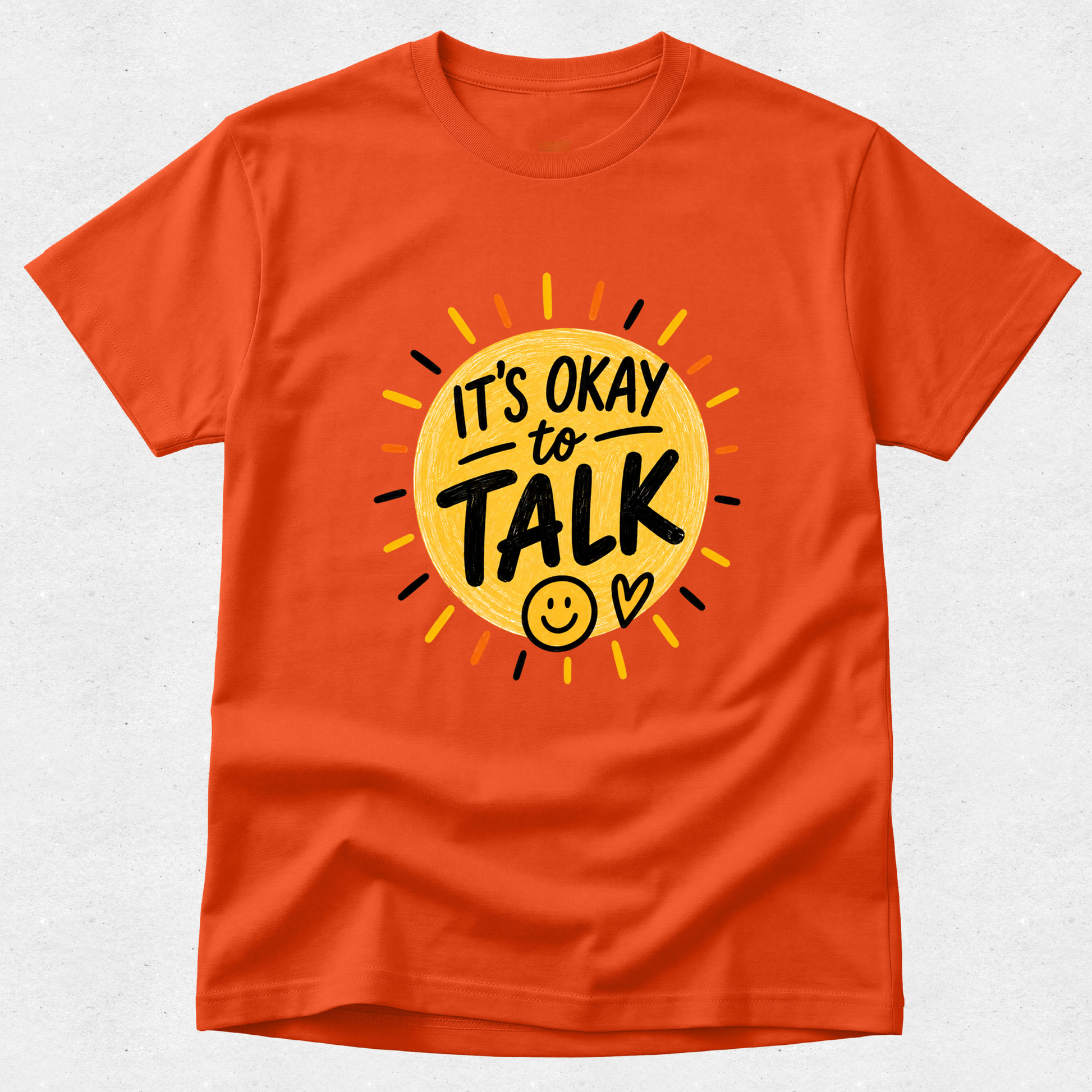 An orange short-sleeve crewneck t-shirt featuring a graphic design of a yellow sun with the text 'IT'S OKAY to TALK' and a smiley face, displayed on a light gray textured background.