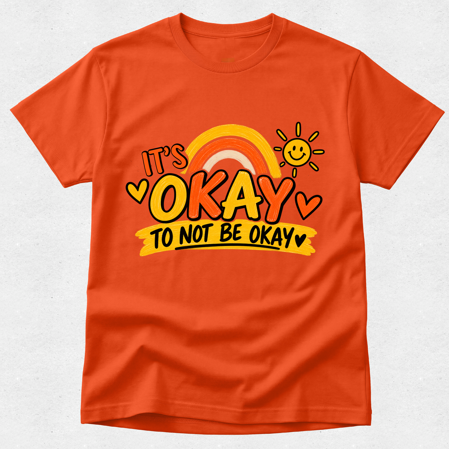 An orange short-sleeve crewneck t-shirt featuring a colorful graphic on the front that reads 'IT'S OKAY TO NOT BE OKAY' with a rainbow, a smiling sun, and hearts, displayed on a light gray textured background.
