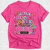A bright pink short-sleeve t-shirt with a colorful graphic print that reads 'THIS IS MY BOOK WEEK COSTUME!' surrounded by illustrations of a crown, wand, makeup palette, sunglasses, and high heels.
