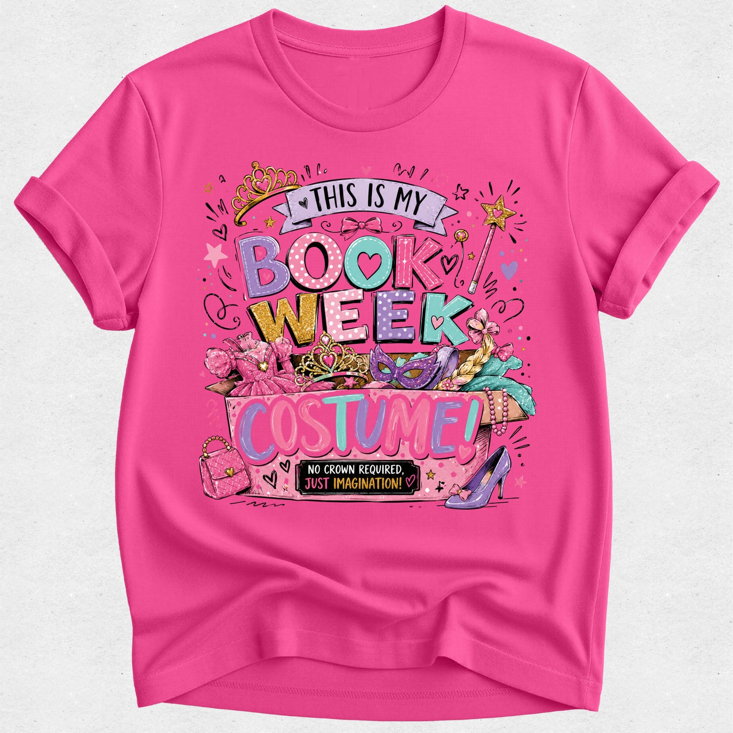 A bright pink short-sleeve t-shirt with a colorful graphic print that reads 'THIS IS MY BOOK WEEK COSTUME!' surrounded by illustrations of a crown, wand, makeup palette, sunglasses, and high heels.