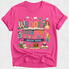 A bright pink short-sleeve t-shirt with a colorful cartoon graphic on the front. The graphic features the words 'BOOK WANDERER ADVENTURES BEGIN HERE' surrounded by whimsical illustrations of a castle, an open book, a rocket, a wizard, a princess, a mermaid, a fairy, a treasure chest, and a fox.