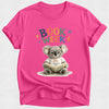A bright pink short-sleeve t-shirt with a 'Book Week' graphic featuring a cartoon koala wearing glasses and reading a book, displayed on a light gray textured background.