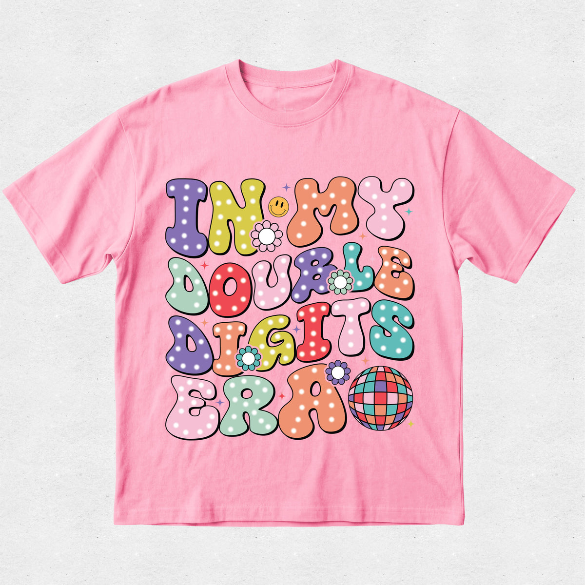 A pink short-sleeve t-shirt with a colorful retro-style graphic on the front that reads 'IN MY DOUBLE DIGITS ERA' in bubbly, polka-dotted letters, displayed flat against a light gray textured background.