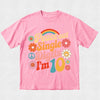 A pink short-sleeve t-shirt with a retro 1960s-style graphic that reads 'Peace out Single Digits I'm 10' in colorful retro lettering, decorated with flowers, a peace sign, and a rainbow, shown on a light textured background.