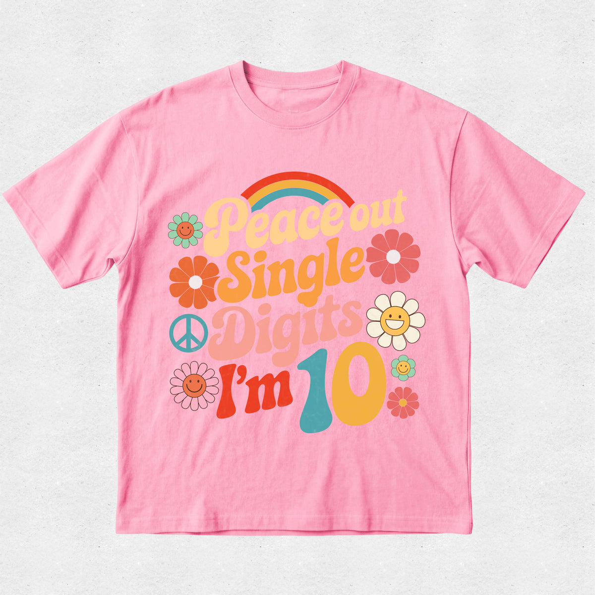 A pink short-sleeve t-shirt with a retro 1960s-style graphic that reads 'Peace out Single Digits I'm 10' in colorful retro lettering, decorated with flowers, a peace sign, and a rainbow, shown on a light textured background.