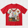 A red short-sleeve crewneck t-shirt featuring a colorful graphic print of a white chef jersey with 'THIS IS MY BOOK WEEK COSTUME!' text and baking-themed illustrations.
