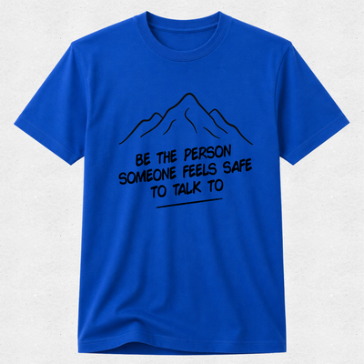A royal blue short-sleeve crewneck t-shirt with a black line art mountain graphic and the text 'BE THE PERSON SOMEONE FEELS SAFE TO TALK TO' printed on the chest, displayed against a light gray textured background.
