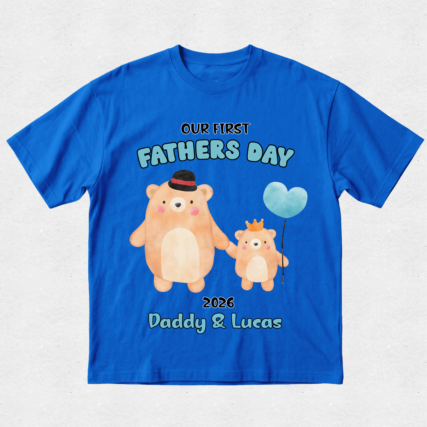 A royal blue short-sleeve t-shirt with a custom Father's Day graphic featuring two cartoon bears holding hands and a light blue heart-shaped balloon. The text reads 'OUR FIRST FATHER'S DAY', '2026', and 'Daddy & Lucas'.