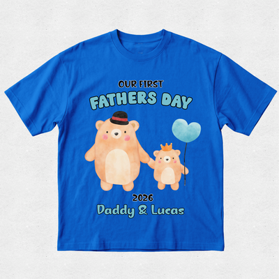 A royal blue short-sleeve t-shirt with a custom Father's Day graphic featuring two cartoon bears holding hands and a light blue heart-shaped balloon. The text reads 'OUR FIRST FATHER'S DAY', '2026', and 'Daddy & Lucas'.