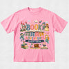 A pink short-sleeve crewneck t-shirt featuring a colorful graphic design on the front that reads 'BOOK WEEK ADVENTURES BEGIN HERE' surrounded by whimsical illustrations of a castle, books, a mermaid, and fantasy characters, displayed flat against a light textured background.