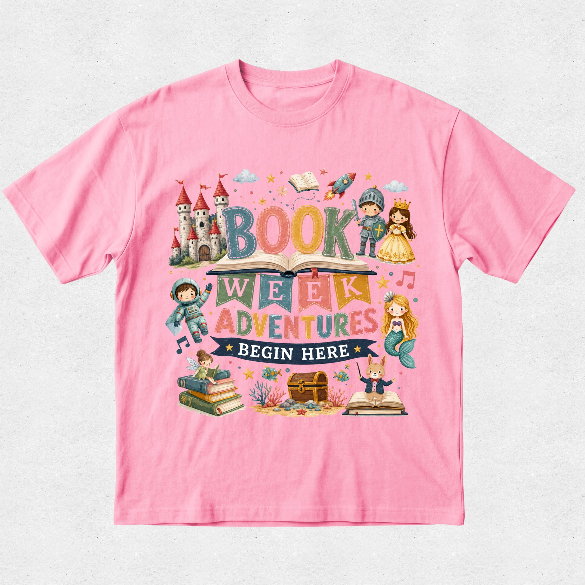 A pink short-sleeve crewneck t-shirt featuring a colorful graphic design on the front that reads 'BOOK WEEK ADVENTURES BEGIN HERE' surrounded by whimsical illustrations of a castle, books, a mermaid, and fantasy characters, displayed flat against a light textured background.
