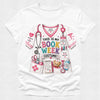 A white short-sleeve crewneck t-shirt featuring a colorful graphic print of a book-themed nurse costume. The graphic includes a pink and white scrub top with a stethoscope, pockets holding a teddy bear and books, and text that reads 'THIS IS MY BOOK WEEK COSTUME!'.