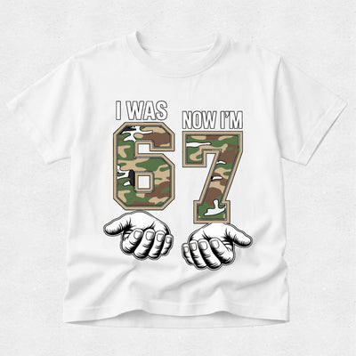 White t-shirt with camouflage numbers '67' and fists, text 'I was 67 now I'm', on a white background.