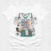 A white short-sleeve crewneck t-shirt featuring a colorful graphic print of a bookish nurse costume. The graphic includes teal scrubs, a stethoscope, medical badges, a teddy bear, and the text 'THIS IS MY BOOK WEEK COSTUME!'.