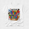 A white short-sleeve crewneck t-shirt featuring a colorful graphic print on the front that reads 'THIS IS MY BOOK WEEK COSTUME!' surrounded by comic-style illustrations of masks, a pirate eye patch, a top hat, a dinosaur, a microphone, and a treasure chest.