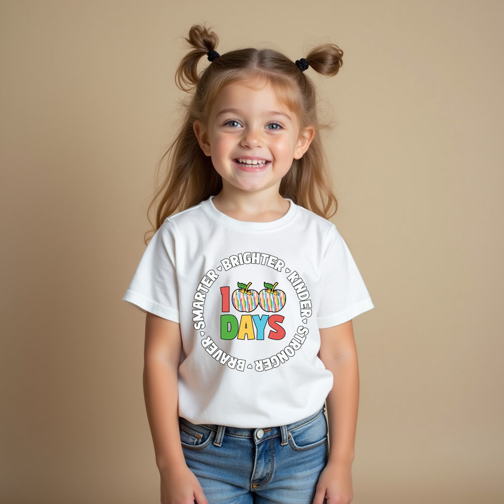 A young girl with blonde pigtails smiles while wearing a white graphic t-shirt and blue jeans against a solid beige studio background. The t-shirt features a colorful circular graphic that reads 'I 100 DAYS SMARTER BRIGHTER WIDER STRONGER BECAUSE' with three small pumpkins and the word 'DAYS' in multicolor letters.