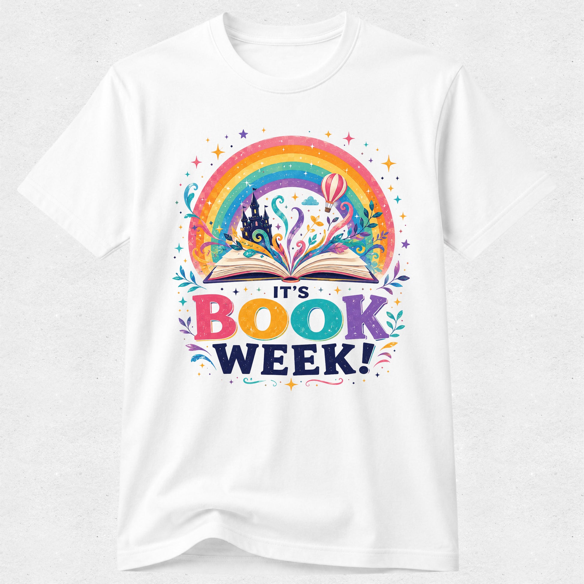 A white short-sleeve crewneck t-shirt featuring a colorful graphic print on the front. The design shows an open book with a rainbow arching over it, a castle silhouette, and the text 'IT'S BOOK WEEK!' in playful multicolored lettering.