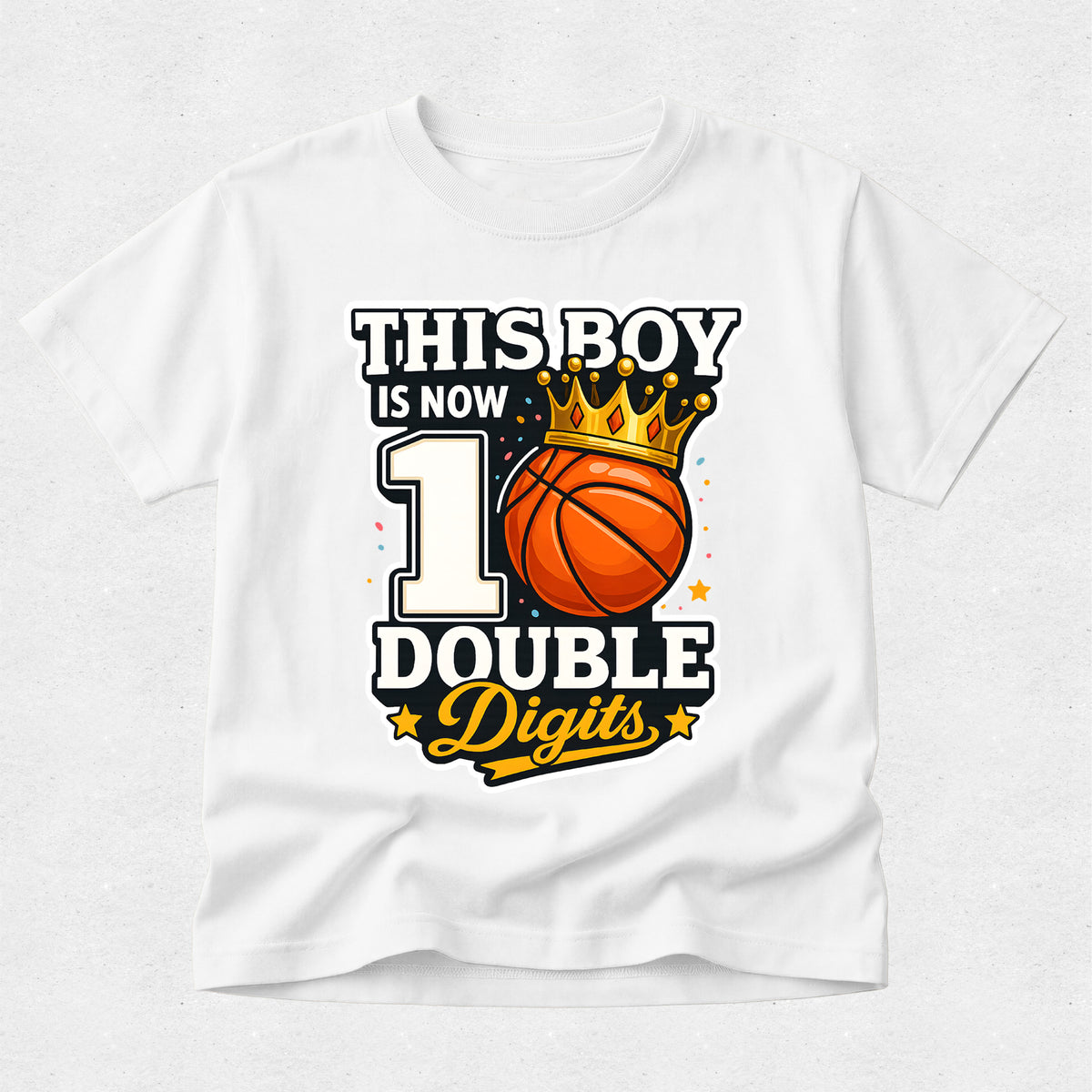 A white short-sleeve t-shirt with a graphic print that reads 'THIS BOY IS NOW 1 DOUBLE Digits' featuring a basketball and a gold crown, displayed on a light gray textured background.
