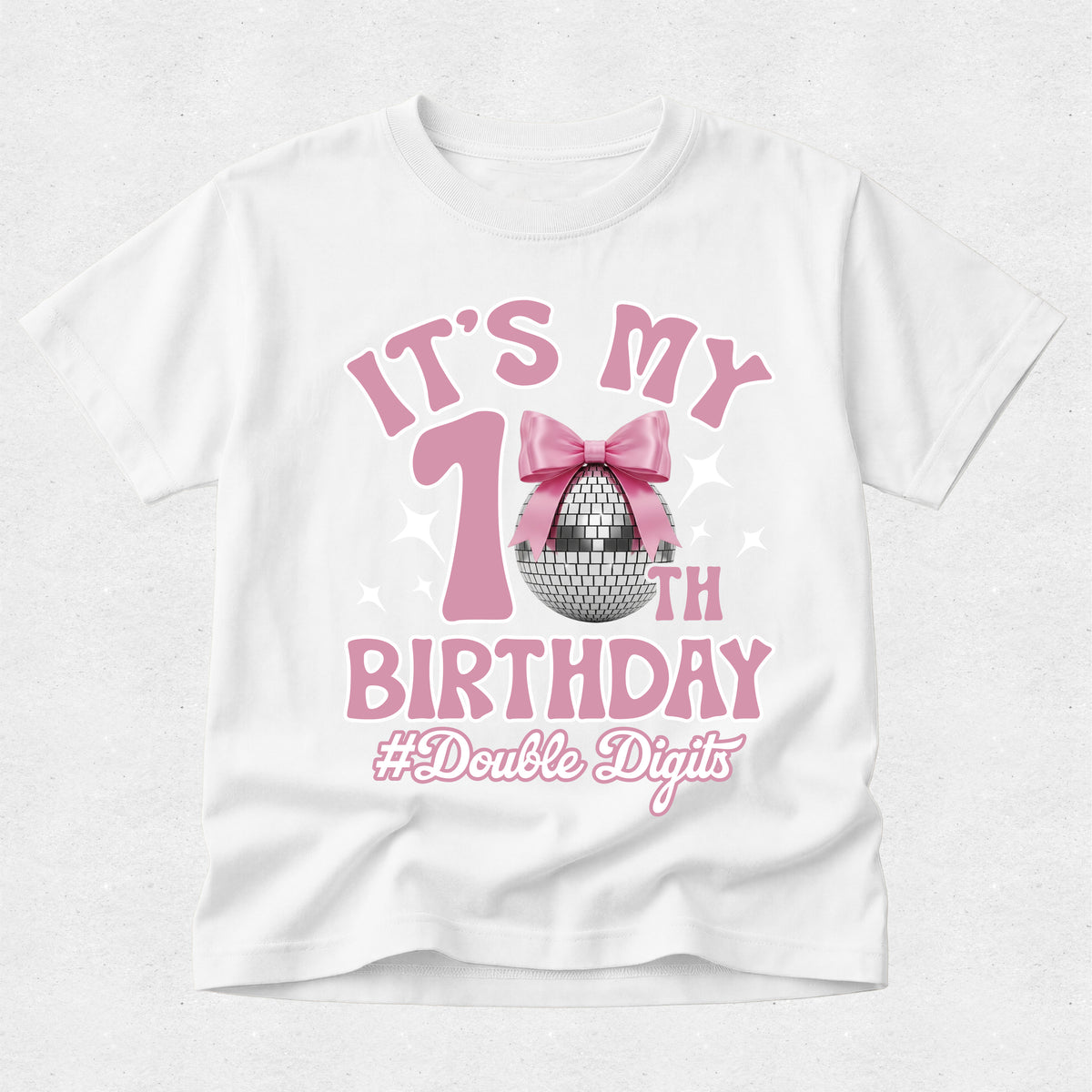 A white short-sleeve t-shirt with a pink and silver graphic that reads 'IT'S MY 1st BIRTHDAY #Double Digits' featuring a pink bow and disco ball design, shown on a light gray textured background.