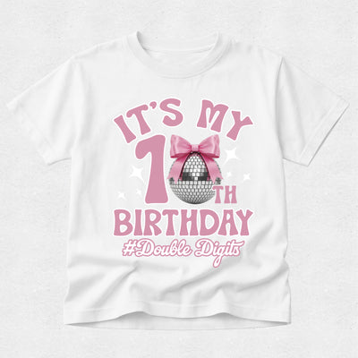 A white short-sleeve t-shirt with a pink and silver graphic that reads 'IT'S MY 1st BIRTHDAY #Double Digits' featuring a pink bow and disco ball design, shown on a light gray textured background.