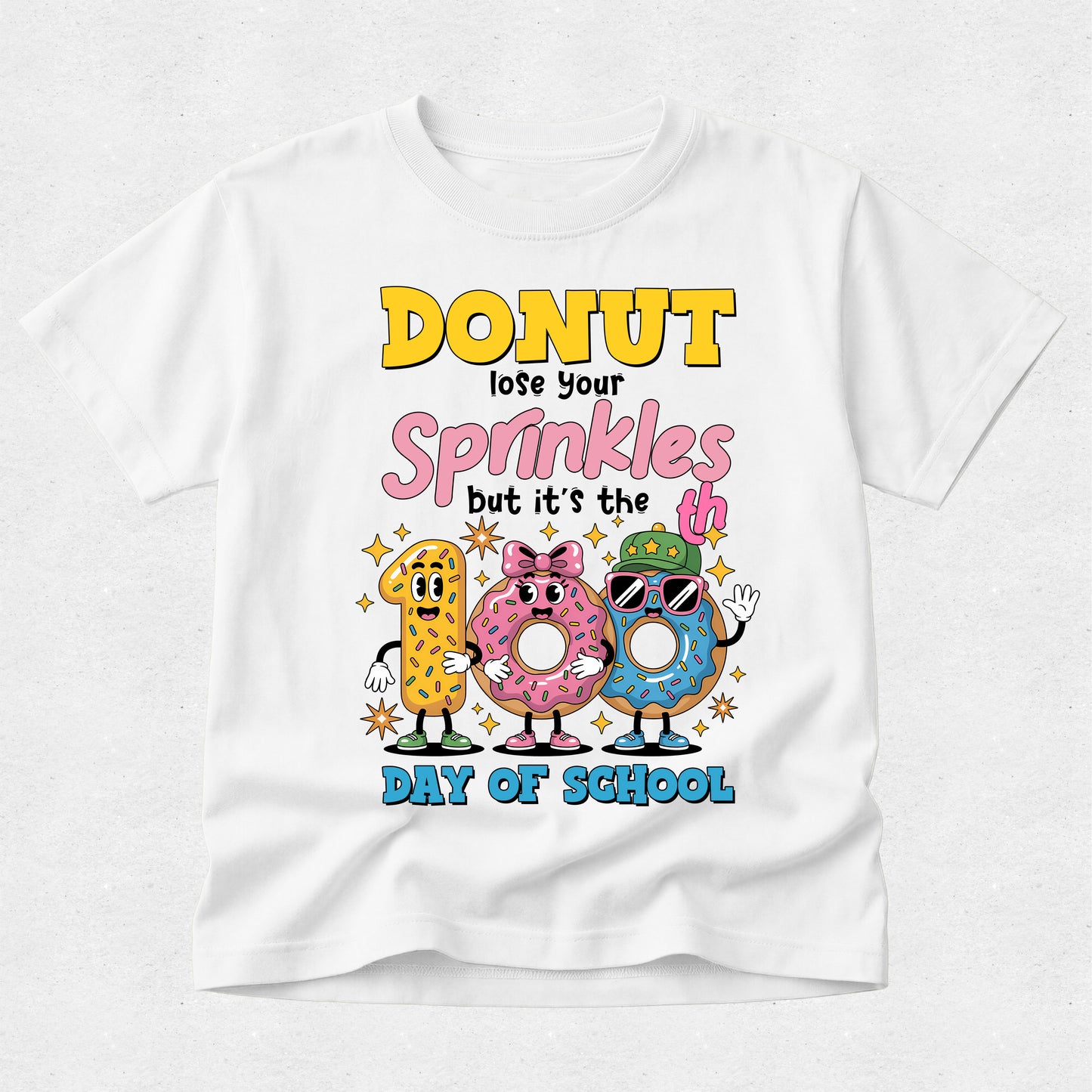A white short-sleeve t-shirt with a colorful graphic on the front that reads 'DONUT lose your Sprinkles but it's the DAY OF SCHOOL' above three cartoon donuts wearing sunglasses and hats, shown on a light gray background.
