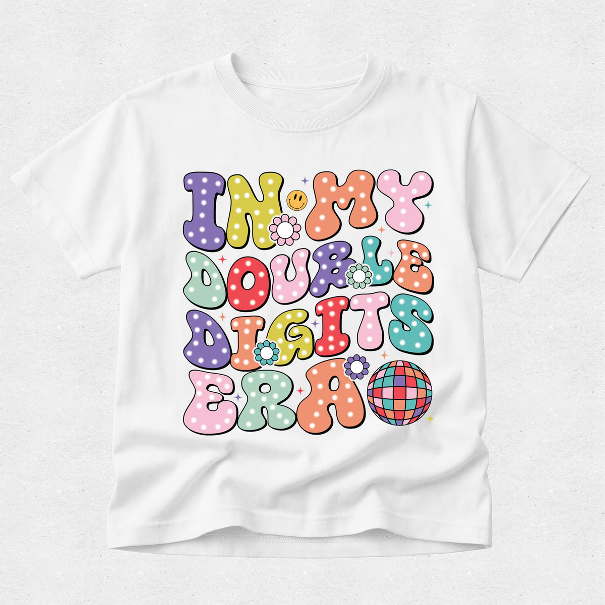 A white short-sleeve crewneck t-shirt featuring a colorful retro graphic that reads 'IN MY DOUBLE DIGITS ERA' in bubbly, polka-dotted letters with a small disco ball motif.