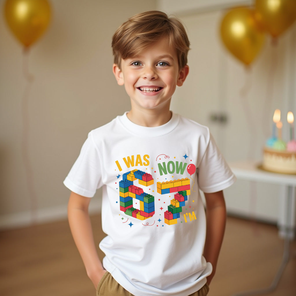 Child wearing a birthday t-shirt with colorful text and building block design, standing in a room with balloons and a cake.
