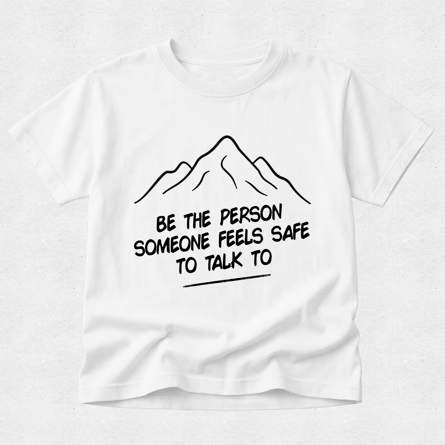 A white short-sleeve t-shirt with a black line drawing of a mountain and the text 'BE THE PERSON SOMEONE FEELS SAFE TO TALK TO' printed on the front, displayed against a light gray textured background.