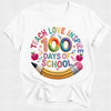 A white short-sleeve crewneck t-shirt featuring a colorful graphic design on the front. The design reads 'TEACH LOVE INSPIRE 100 DAYS OF SCHOOL' in playful, multi-colored lettering, decorated with cartoon apples, a yellow pencil, and small pink flowers.