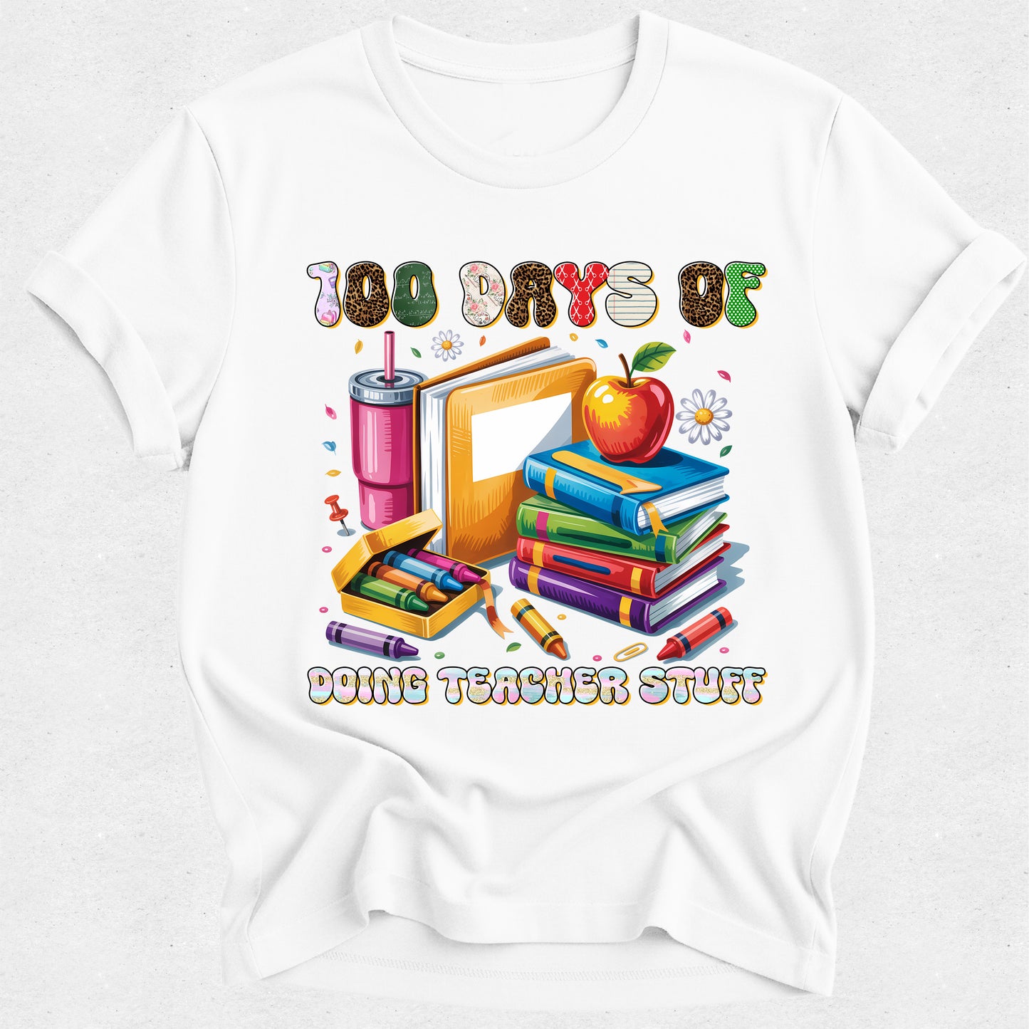A white short-sleeve crewneck t-shirt featuring a colorful graphic print on the front. The graphic displays school supplies including a stack of books, an apple, a pink drink, crayons, and a pencil, with the text '100 DAYS OF DOING TEACHER STUFF' written above and below the illustration.