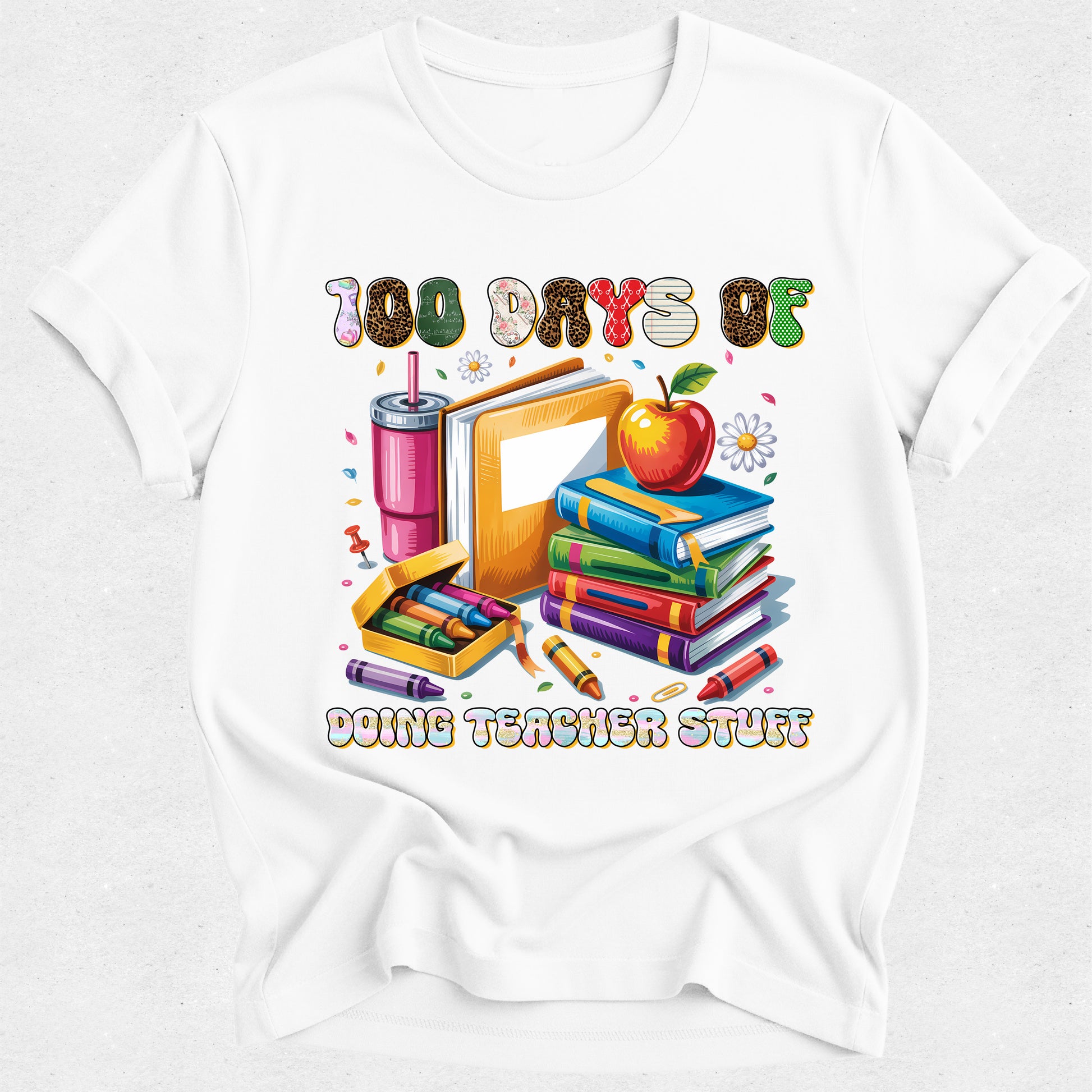 A white short-sleeve crewneck t-shirt featuring a colorful graphic print on the front. The graphic displays school supplies including a stack of books, an apple, a pink drink, crayons, and a pencil, with the text '100 DAYS OF DOING TEACHER STUFF' written above and below the illustration.