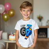 Child wearing a birthday t-shirt with number 6 and 7 design, standing in a room with balloons and cake.