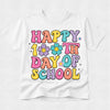 A white short-sleeve t-shirt with a colorful retro-style graphic that reads 'HAPPY 100TH DAY OF SCHOOL' in pastel pink, yellow, blue, purple, and orange bubble letters with flower and star accents, shown on a light gray background.
