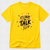 A bright yellow short-sleeve t-shirt with a graphic on the chest that reads 'IT'S OKAY to TALK' in black lettering inside a sunburst design with small orange, red, and black rays, shown on a light gray textured background.