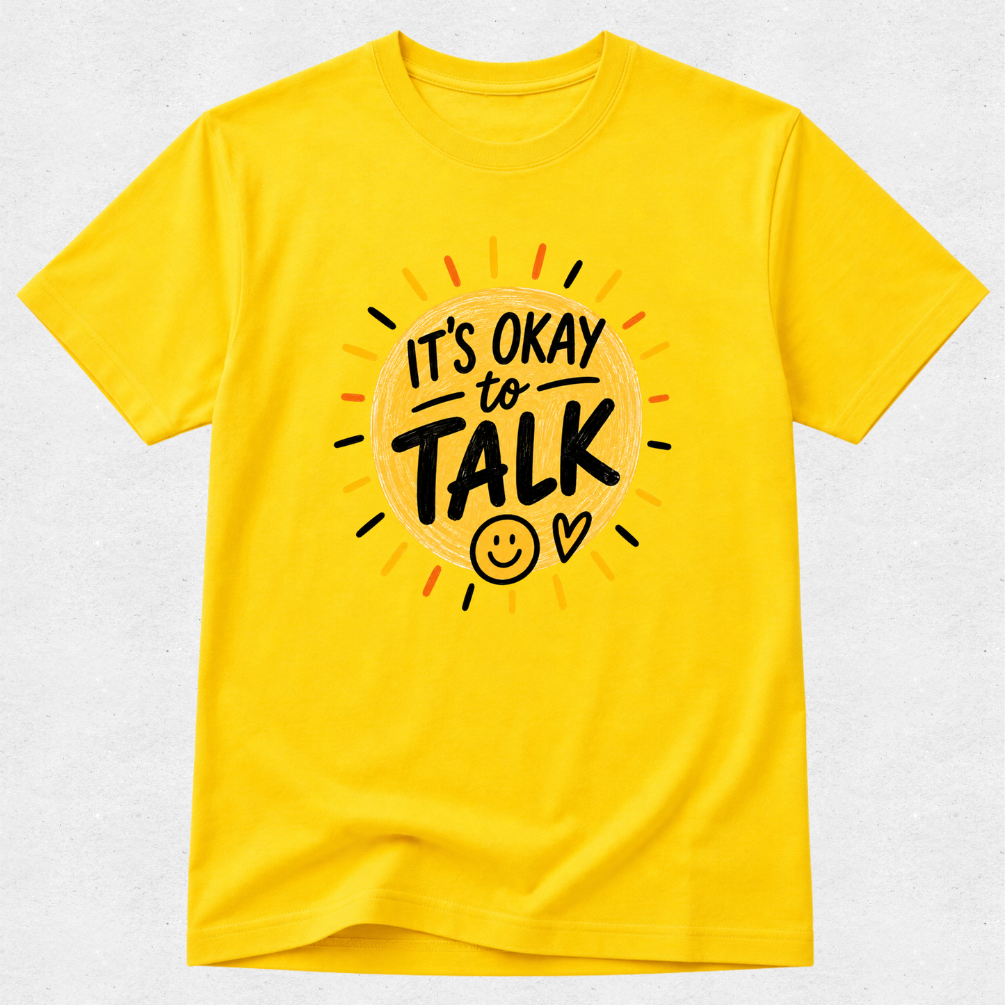 A bright yellow short-sleeve t-shirt with a graphic on the chest that reads 'IT'S OKAY to TALK' in black lettering inside a sunburst design with small orange, red, and black rays, shown on a light gray textured background.