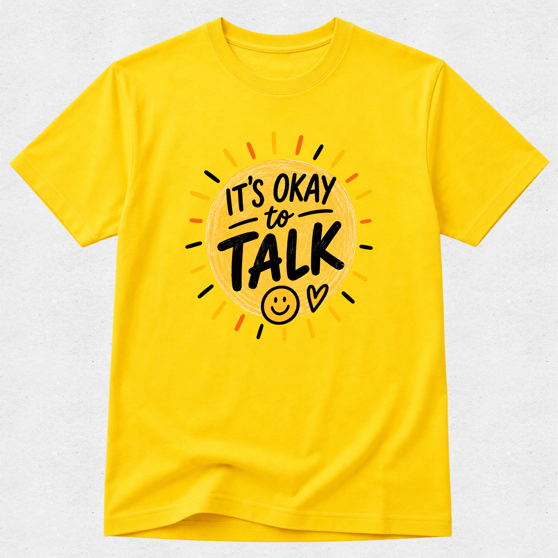 A bright yellow short-sleeve t-shirt with a graphic on the chest that reads 'IT'S OKAY to TALK' in black lettering inside a sunburst design with small orange, red, and black rays, shown on a light gray textured background.