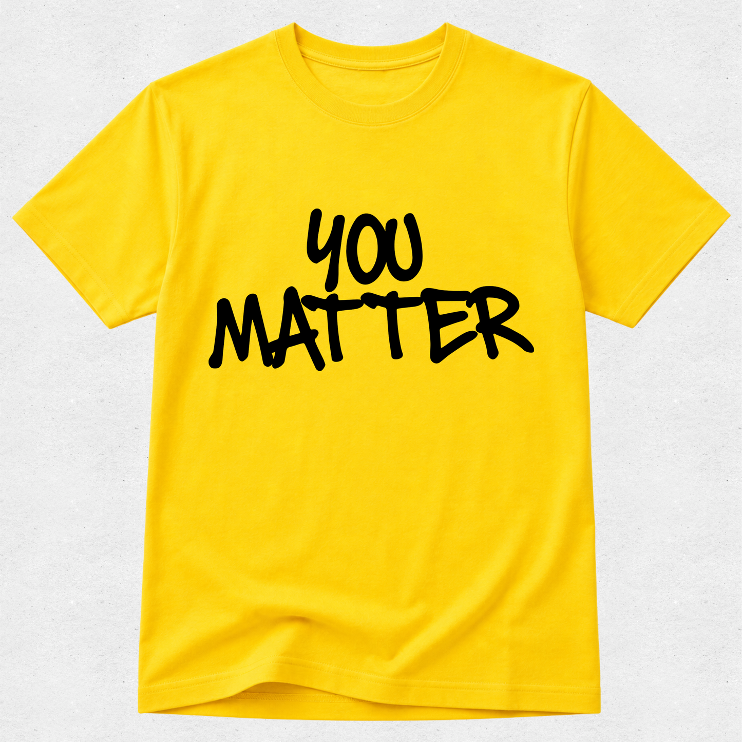 A bright yellow short-sleeve t-shirt with the words 'YOU MATTER' printed across the chest in a black handwritten-style font, displayed against a light gray textured background.