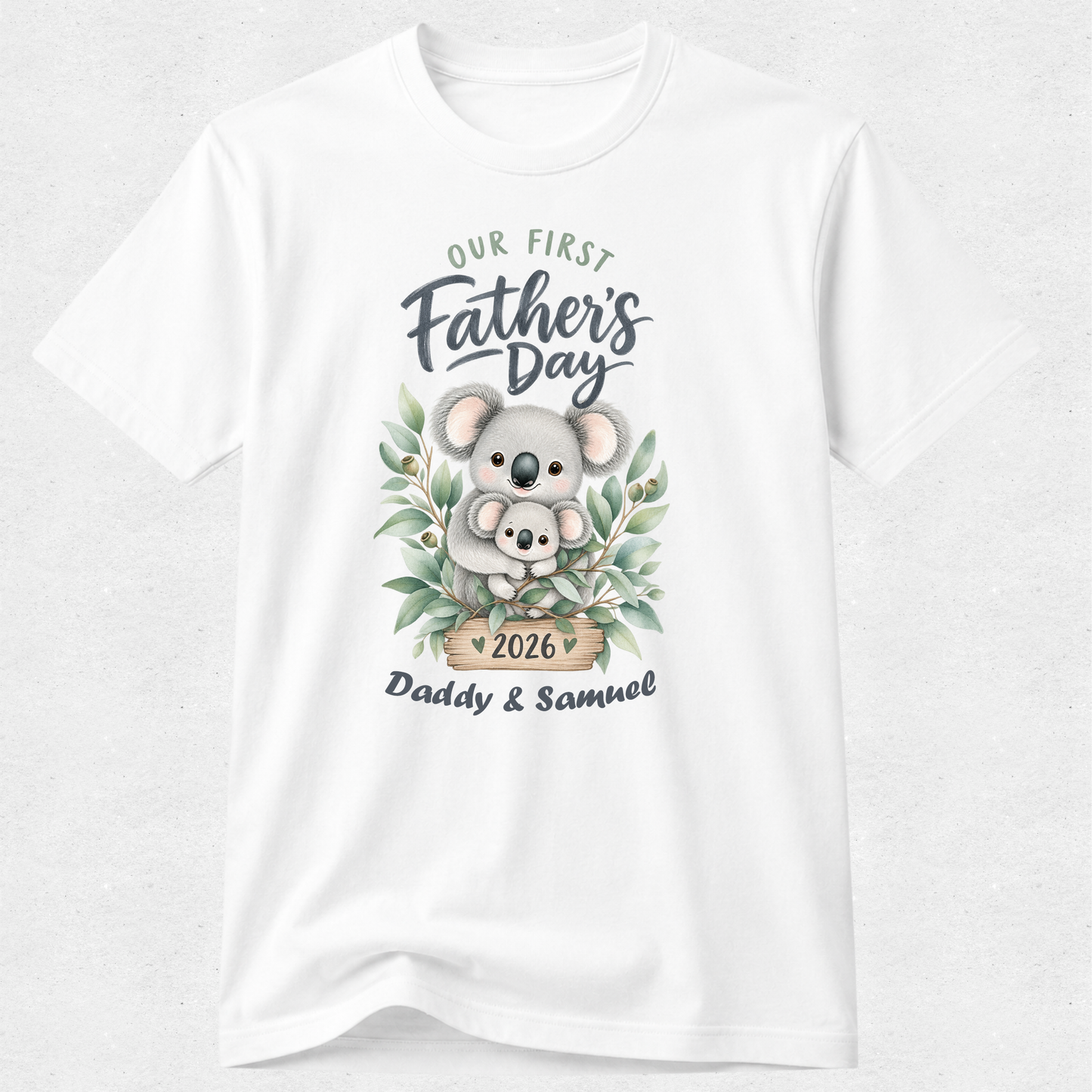 A white short-sleeve crewneck t-shirt featuring a watercolor-style graphic of a koala holding a baby koala, surrounded by green leaves. The text reads 'Our First Father's Day' and 'Daddy & Samuel 2026'.