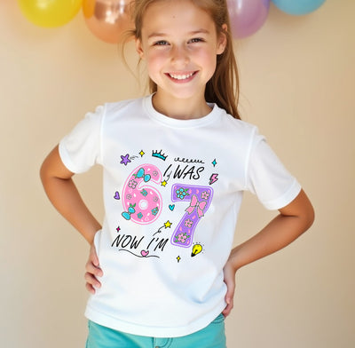 Young girl wearing a white t-shirt with colorful text 6 7 and numbers, standing against a beige background with balloons.