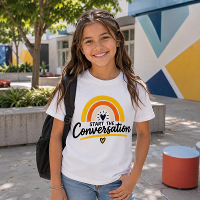 A smiling young woman standing outdoors in a sunny plaza, wearing a white graphic T-shirt with a rainbow and the words 'START THE Conversation' printed on the front, along with blue jeans and a black backpack.