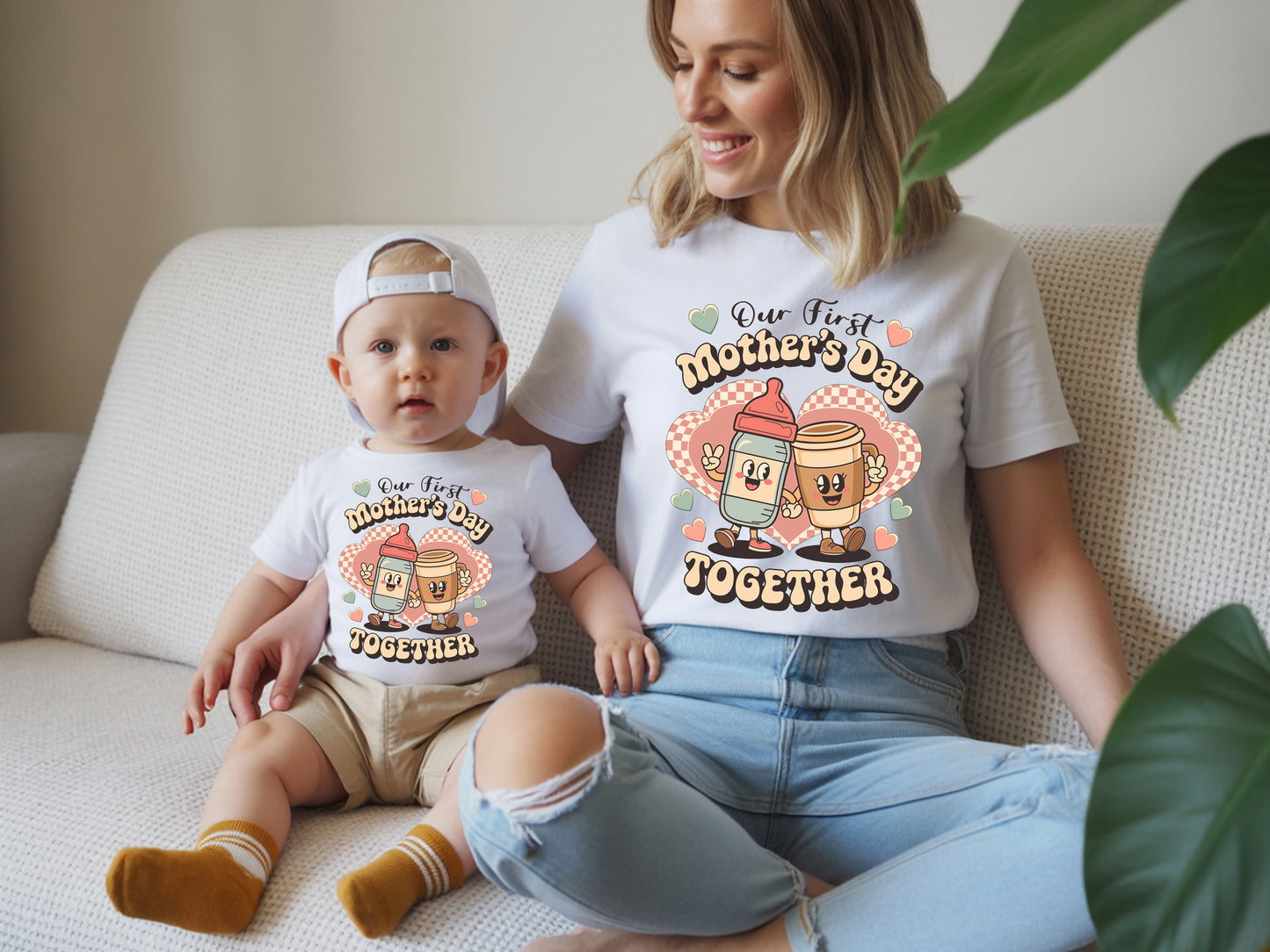 Woman and baby sitting on a couch wearing matching 'First Mother's Day Together' shirts.