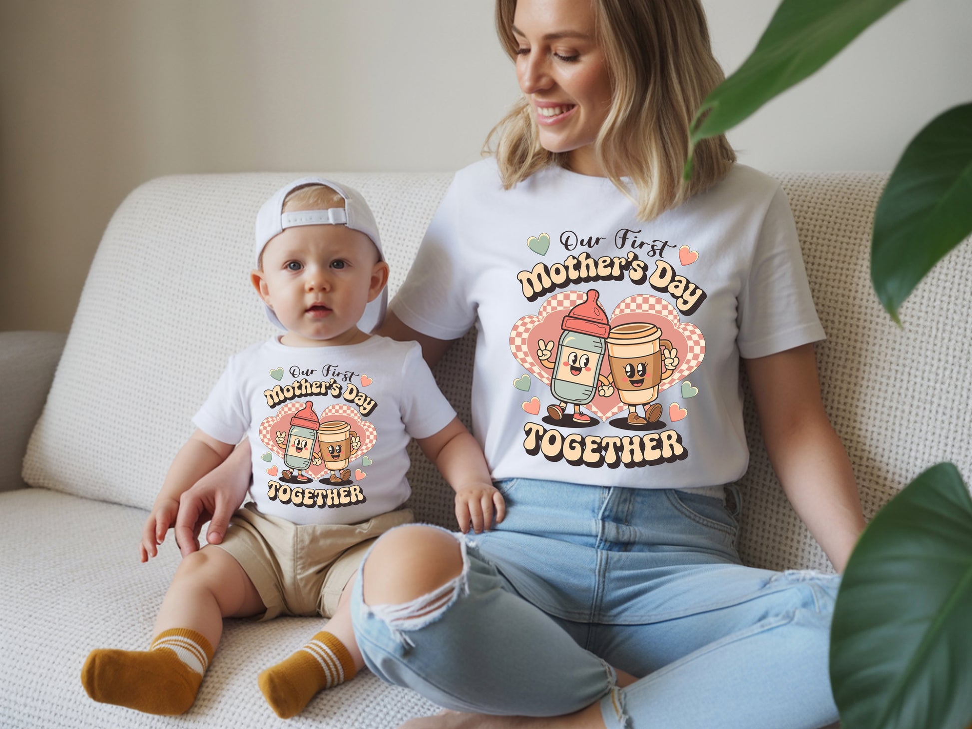 Woman and baby sitting on a couch wearing matching 'First Mother's Day Together' shirts.