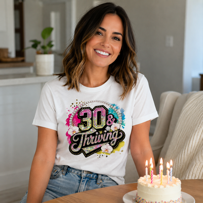 Woman wearing a white t-shirt with '30 & Thriving' design, sitting next to a birthday cake with lit candles.