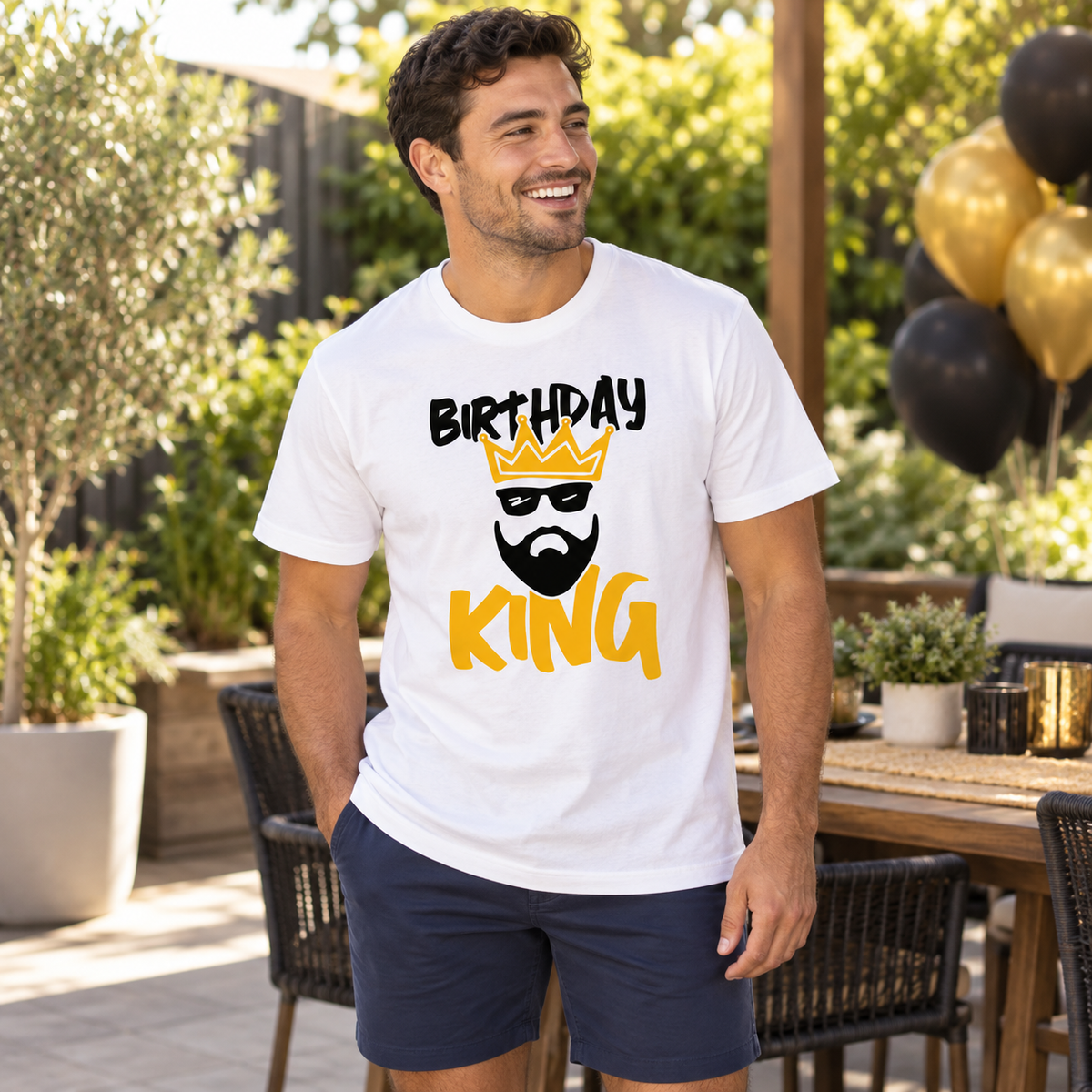 A man wearing a white graphic t-shirt with a 'BIRTHDAY KING' design featuring a crown and beard, standing in a sunny outdoor patio setting.
