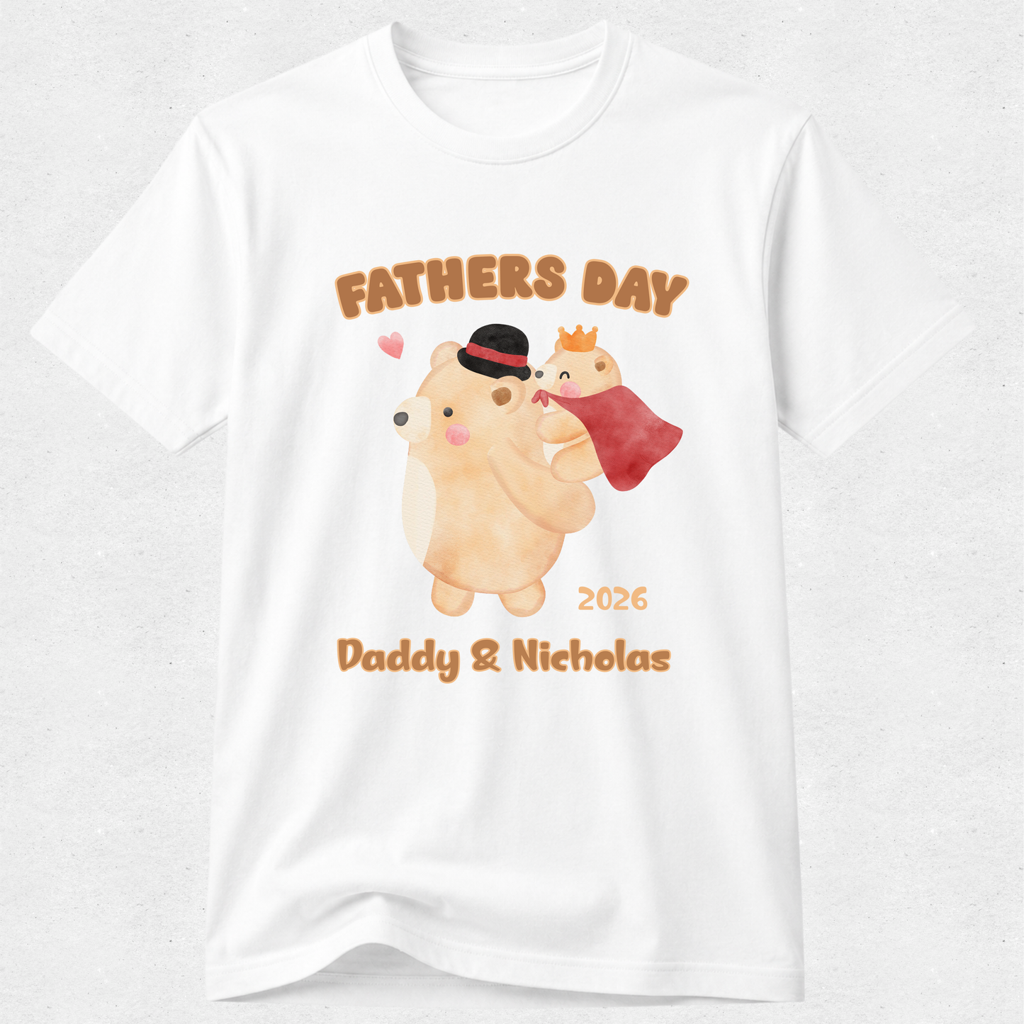 A white short-sleeve crewneck t-shirt featuring a custom watercolor-style graphic of two bears on a light gray background. The graphic includes the text 'FATHERS DAY' in brown, a watercolor illustration of a tan bear wearing a black top hat and holding a red stocking, a small crowned bear, '2026', and 'Daddy & Nicholas' in brown.