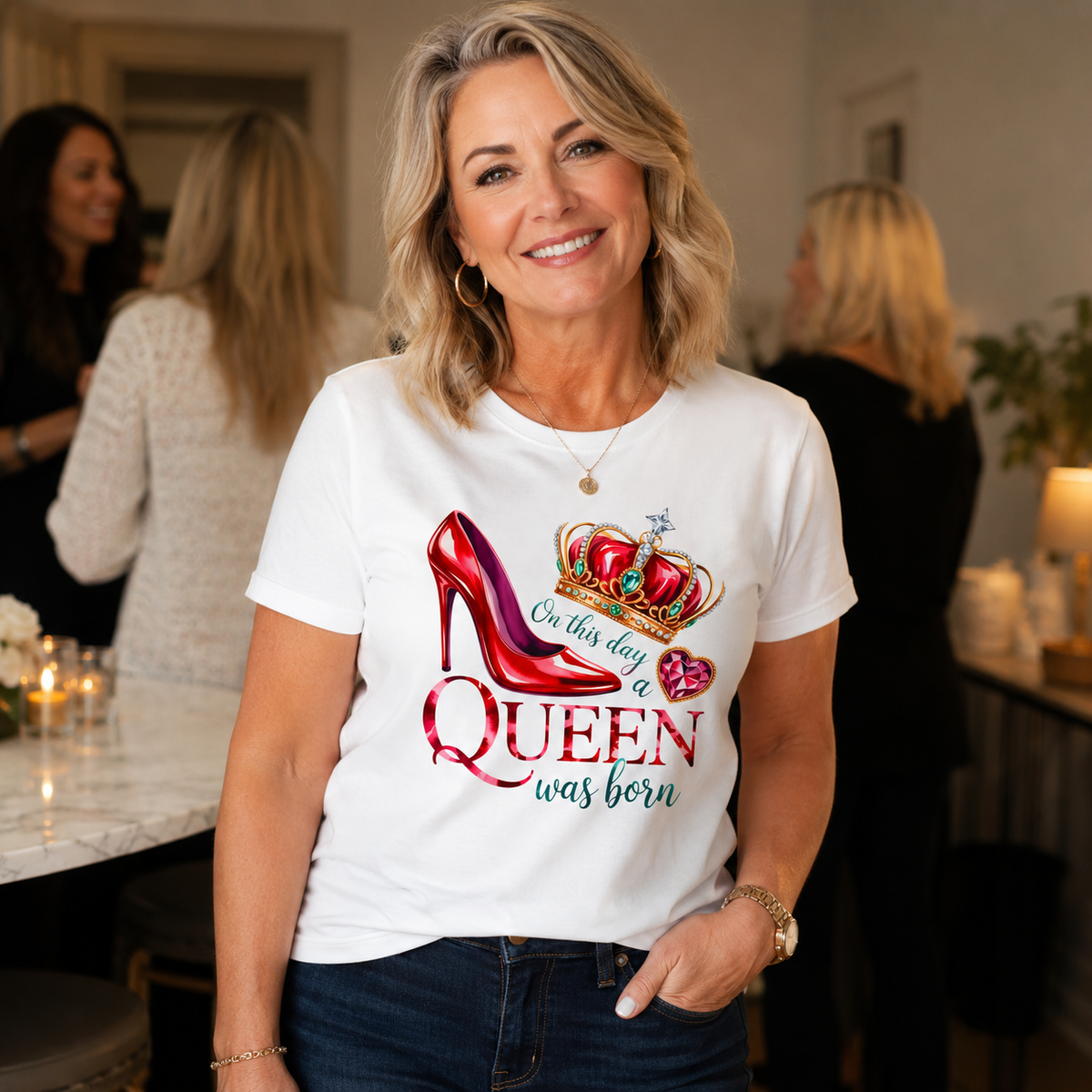 A smiling blonde woman wearing a white graphic t-shirt with a red high-heel shoe, crown, and the text 'On this day a QUEEN was born' in a social event setting.
