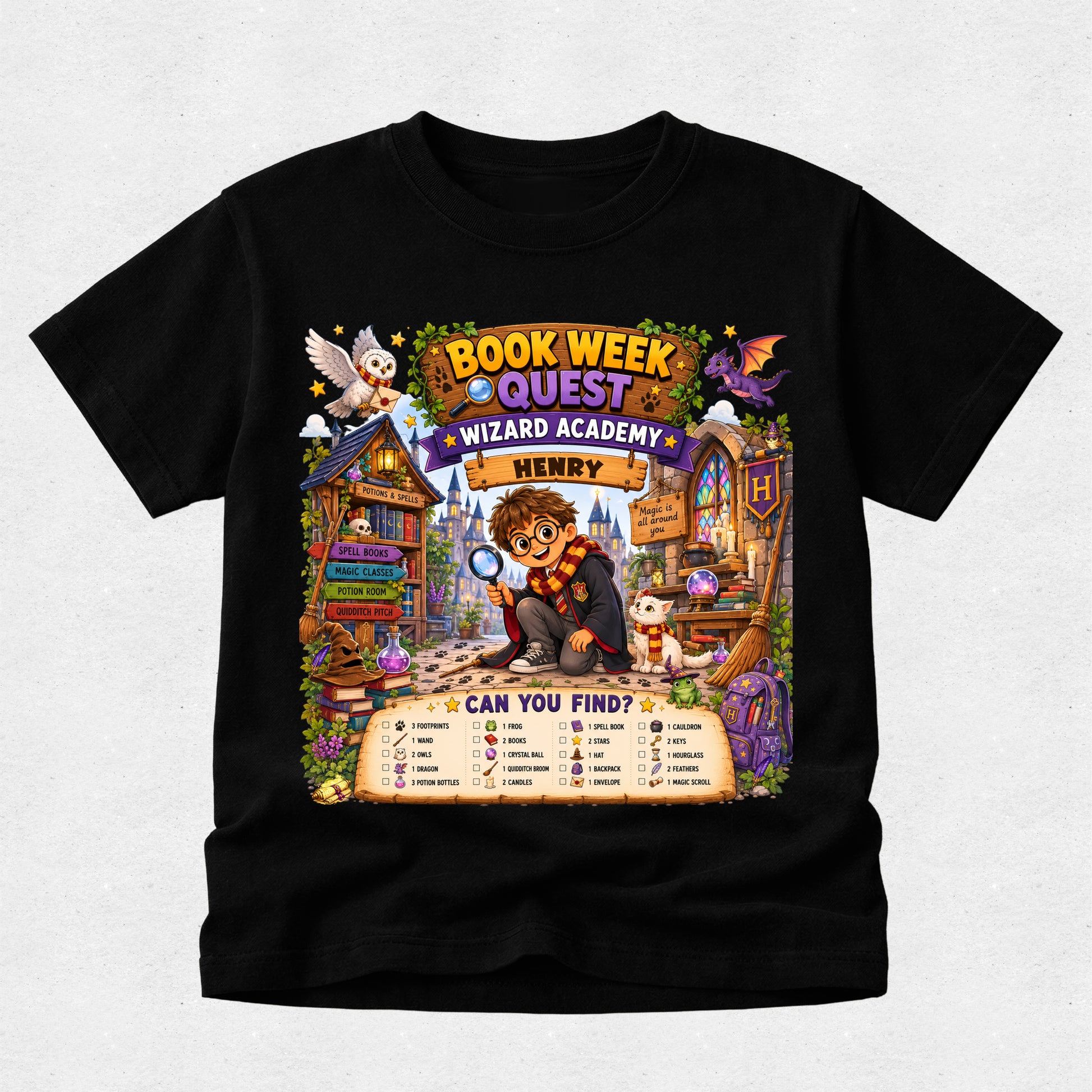 A black short-sleeve crewneck t-shirt featuring a colorful graphic print for 'Book Week' at 'Wizard Academy' about 'Henry'. The graphic shows a cartoon lion in a wizard outfit, a cat, a owl, a dragon, and a 'Can You Find?' word search grid at the bottom, displayed on a light gray textured background.